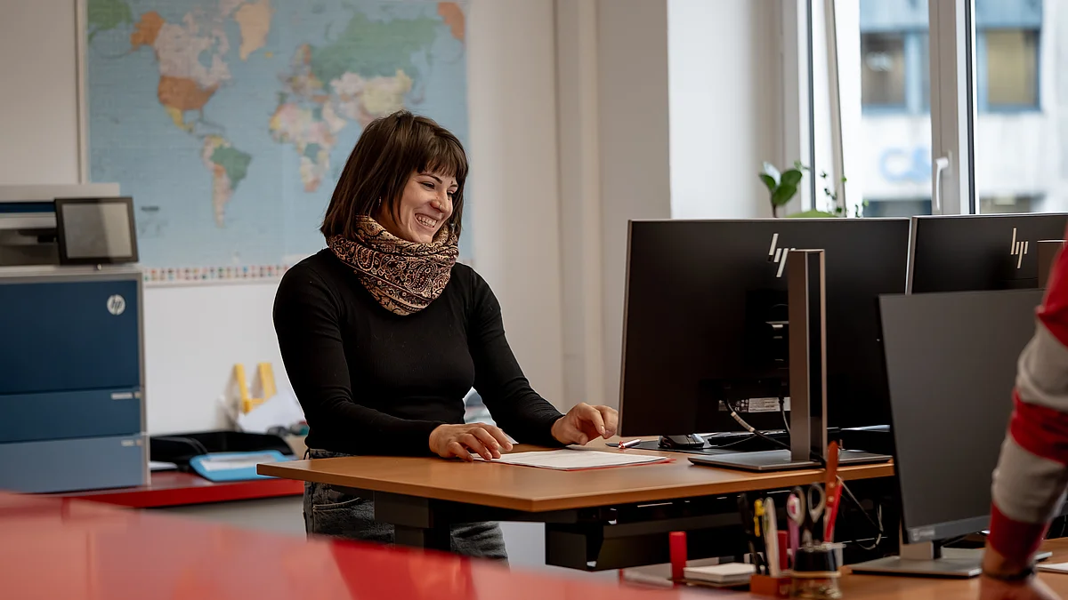 Eine Frau sitzt lächelnd an einem Schreibtisch im Büro, arbeitet an einem Dokument und blickt auf ihren Computerbildschirm.