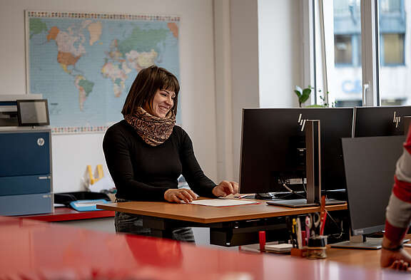 Eine Frau sitzt lächelnd an einem Schreibtisch im Büro, arbeitet an einem Dokument und blickt auf ihren Computerbildschirm.