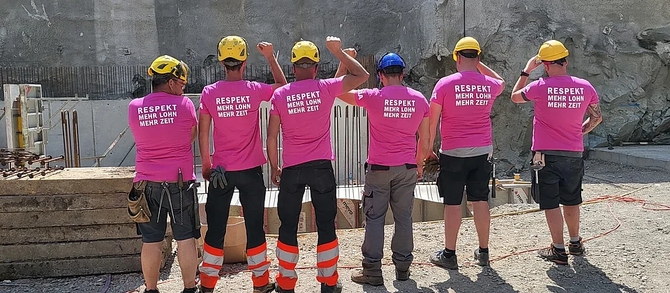 Sechs Bauarbeiter auf einer Baustelle tragen pinke T-Shirts der Gewerkschaft Unia mit der Aufschrift «Respekt, mehr Lohn, mehr Zeit» und gelbe oder blaue Schutzhelme.